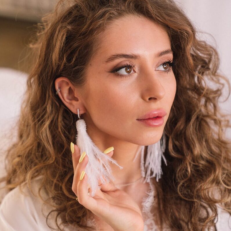 Portrait of a young woman with curly hair wearing elegant feather earrings indoors.